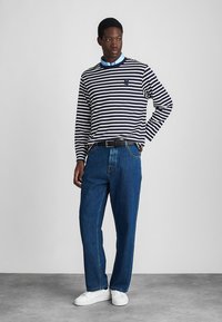 Mann in navy- und weißen gestreiften Langarmshirt über einem blauen Hemd, blaue Jeans, schwarzem Gürtel und weißen Turnschuhen, der vor einem schlichten Hintergrund steht.