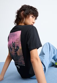 Black cotton t-shirt with a colorful graphic print on the back, featuring a cartoon character against a pink sky and urban silhouette.