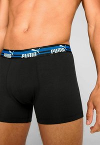 Black boxer shorts with a blue and white striped elastic waistband featuring “PUMA” branding. Fabric appears smooth and stretchy.