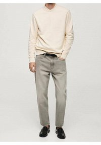 Mężczyzna w beżowej bluzie, jasnoszarych dżinsach z czarnym paskiem i czarnych skórzanych mokasynach, stojący z jedną ręką w kieszeni.