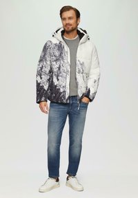 Mann in einer weiß-schwarzen, bergmusterigen Steppjacke, grauem Pullover, blauen Jeans und weißen Sneakers, der mit einer Hand in der Tasche steht.