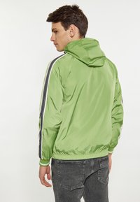 Giacca leggera verde con cappuccio, caratterizzata da strisce verticali bianche e nere lungo le maniche e un orlo fitted. Finitura texturizzata e lucida.