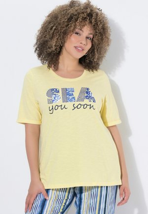 Frau mit lockigem Haar, die ein gelbes Shirt mit dem Schriftzug "SEA you soon" und gestreifte blaue, gelbe und weiße Hosen trägt, posiert vor einem weißen Hintergrund.