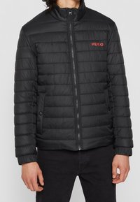 Czarna kurtka puchowa z wysokim kołnierzem, pikowaną teksturą, zamek błyskawiczny z przodu oraz czerwonym logo "HUGO" na piersi. Zawiera boczne kieszenie z zatrzaskami.