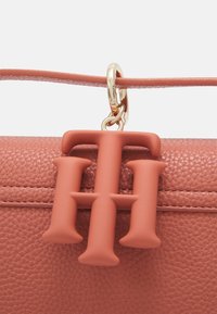 Bolso de mano de cuero color coral con un acabado texturizado, que presenta un destacado charm con logo en tono durazno mate y herrajes en tono dorado. Detalle de diseño elegante.