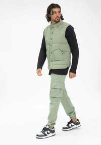 Gilet matelassé vert avec fermeture à boutons et poches frontales, associé à un pantalon cargo vert clair et des baskets noir et blanc.