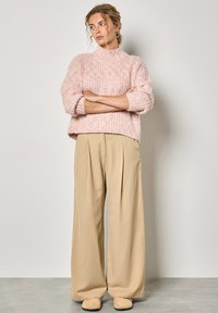 Jasnoróżowy sweter z dzianiny z wysokim dekoltem, w połączeniu z beżowymi spodniami o szerokich nogawkach. Stroje wyróżniają się luźnym krojem i teksturowanym materiałem.
