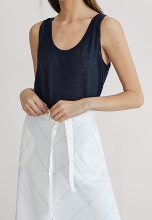 Kvinde iført ærmeløs navyblå top stoppet ned i en hvid nederdel med høj talje og diskret diagonal syning, binder et bælte i taljen.