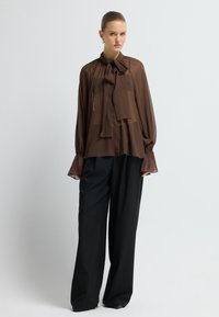 Frau in einer durchsichtigen braunen Bluse mit Schleife und weiten Ärmeln, kombiniert mit lockeren schwarzen weiten Hosen, steht vor einem einfarbigen Hintergrund.