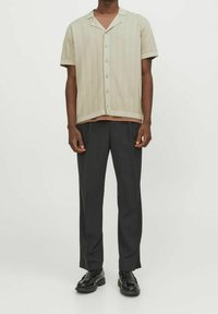 Man som bär en beige kortärmad knäpp kardigan över en brun skjorta, svarta byxor och svarta läderskor mot vit bakgrund.