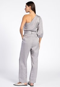 Zilveren, een-schouder jumpsuit van textuurstof met een pofmouw. De brede broekspijpen hebben een ceintuur in de taille en schuine zakken.