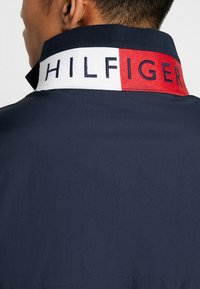 Mand iført en marineblå skjorte med opadvendt krave, der har en hvid og rød panel broderet med "HILFIGER" i marineblå bogstaver.