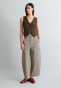 Braunes, geripptes ärmelloses Weste mit Knopfdetail, kombiniert mit weit geschnittenen karierten Hosen in Erdtönen und roten Lederschuhen.