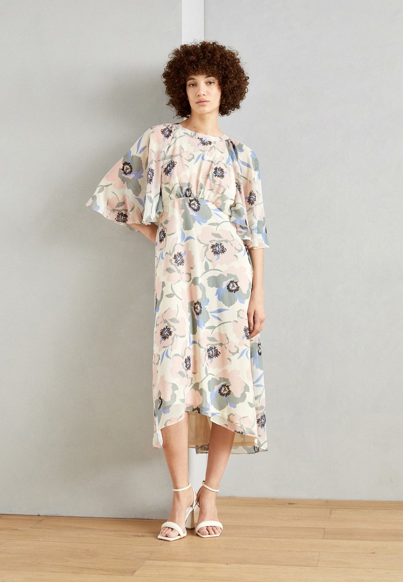 Robe florale mi-longue avec des manches volantées, présentant des couleurs pastel et des motifs floraux sombres. Accompagnée de sandales blanches à lanières.