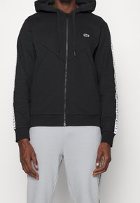 Svart zip-up hoodie med kängurufickor, strukturerad tyg, mjukt innerfoder, med en grön logotyp och vita sidostråk.