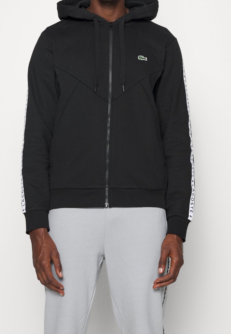 Svart zip-up hoodie med kängurufickor, strukturerad tyg, mjukt innerfoder, med en grön logotyp och vita sidostråk.