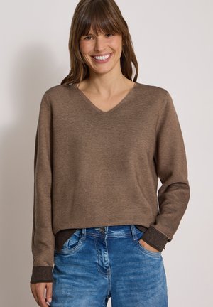 Femme souriante portant un pull à col en V marron rayé et un jean bleu, les mains partiellement dans les poches, sur un fond uni.
