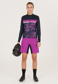 Tät passform, långärmad cykeltröja i navy och rosa med ett mönstrat framstycke. Matchande ljusrosa shorts med svarta accenter. Hjälm i handen.