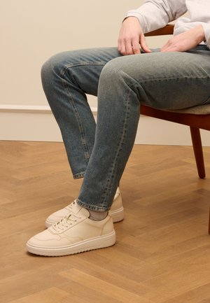 Sneakers in pelle beige con punta arrotondata, suola piatta e dettagli cuciti, abbinati a jeans blu sbiaditi, seduti su una sedia di legno.