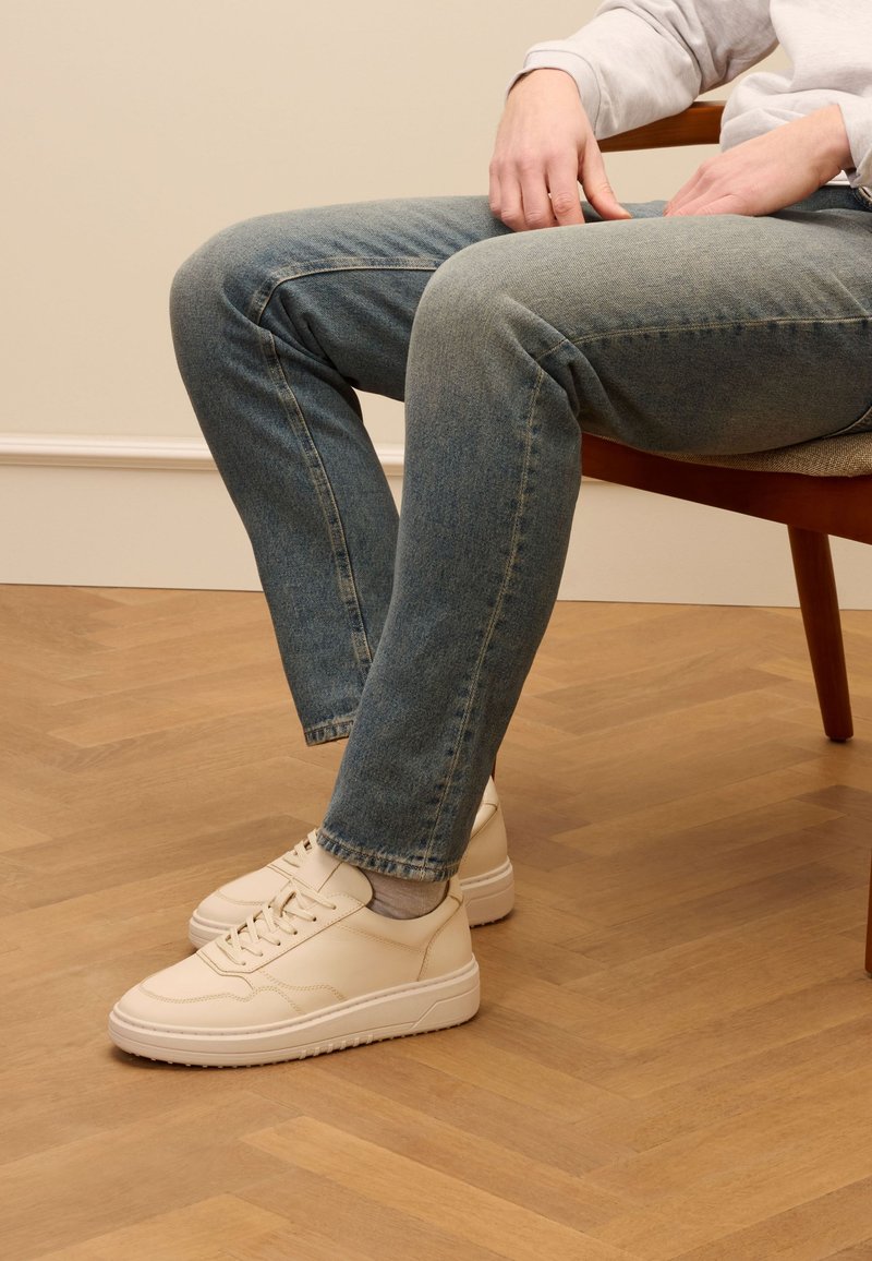 Sneakers in pelle beige con punta arrotondata, suola piatta e dettagli cuciti, abbinati a jeans blu sbiaditi, seduti su una sedia di legno.