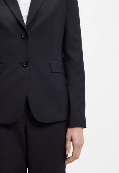 Blazer noir cintré avec deux boutons et poches à rabat, porté avec un pantalon assorti, visible de la taille aux mains sur fond blanc.