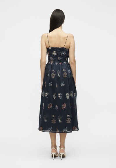 Femme aux longs cheveux foncés portant une robe midi bleu marine à fines bretelles avec un motif floral abstrait multicolore, associée à des chaussures blanches à talons.