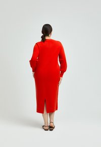 Rotes Strickkleid mit langen Puffärmeln, einem Schlitz im Rücken und einem runden Ausschnitt. Glatte Textur und figurbetonte Form bis zur Mitte der Wade.