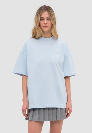 Young woman wearing a light blue oversized T-shirt and a gray pleated mini skirt, standing against a plain background.