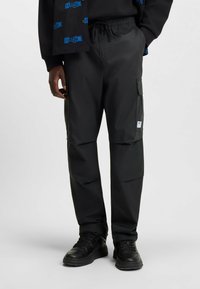 Zwarte cargobroek van gladde stof, met een trekkoord in de taille en zijzakken. Gecombineerd met een zwarte sneaker met een gestructureerd oppervlak.