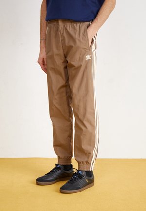 Osoba nosící hnědé tepláky Adidas s bílými pruhy a černé tenisky, stojící na žluté podlaze před jednobarevným pozadím.