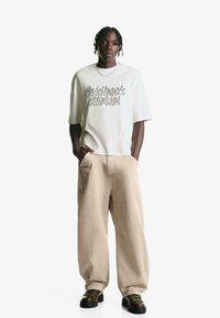 Krátký rukáv krémové tričko s černým kurzívním textem, spárované s volnými béžovými kalhotami a tmavými botami s zelenými tkaničkami.