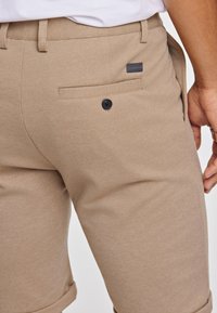 Beżowe szorty wykonane z fakturowanej tkaniny, z tylną kieszenią zapinaną na czarny guzik i małą naszywką z logo. Widoczny podwinięty brzeg.