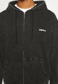 Felpa con cappuccio nera con cerniera realizzata in tessuto morbido. Presenta una tasca frontale, cappuccio con coulisse e un piccolo logo bianco sul petto. Finitura invecchiata.