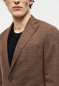 Άνδρας με καφέ σακάκι με υφή πάνω από μαύρο πουκάμισο, που δείχνει το καρφιτσωτό πέτο και τις λεπτομέρειες των κουμπιών, με σκουλαρίκια εμφανή στα αυτιά.