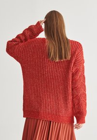 Czerwony sweter z dzianiny o luźnym kroju, z teksturowanym wzorem i warkoczowymi rękawami. Posiada ściągacz na dole oraz konstrukcję z opadającymi ramionami.