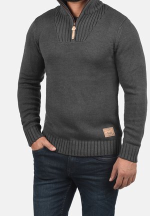 Mann trägt dunkelgrauen gestrickten Pullover mit halbhohem Reißverschlusskragen und Lederpatch auf der Vorderseite, kombiniert mit dunkelblauen Jeans, Hände locker.