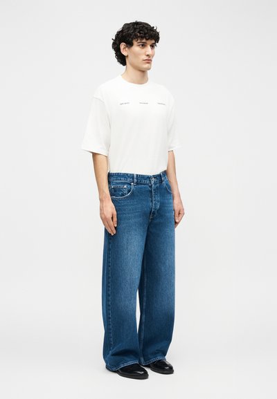 Jeune homme aux cheveux bouclés portant un T-shirt blanc surdimensionné et un jean bleu ample, debout devant un fond clair uni, légèrement tourné vers la droite.