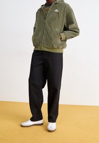 Olivgrön kordflodjacka med dragkedja och sidofickor, i kombination med lösa svarta byxor och vita sneakers. Bakgrunden är i mjuk gul.