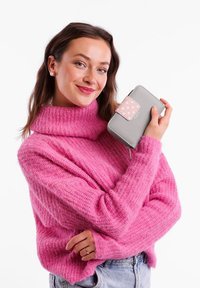 Gray wallet with pink polka dot accent, zip closure. Model in pink fuzzy sweater and blue jeans holds wallet against a white background.