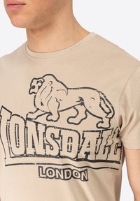 Bež majica od pamuka s velikim crnim grafičkim prikazom lava i tekstom "Lonsdale". Okrugli izrez i kratki rukavi s glatkom teksturom.