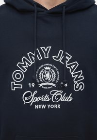 Sweat à capuche bleu marine avec texte blanc brodé "Tommy Jeans Sports Club New York" et noms de villes entourant un emblème de blason de lion.