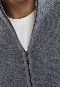 Záběr zblízka na osobu oblečenou v šedém pleteném svršku na zip přes bílý tričko s kulatým krkem. Zip je částečně rozepnutý.