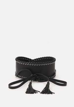 Ceinture large noire avec œillets argentés le long des bords et deux pompons noués à l'avant sur des lanières en cuir sur fond blanc.