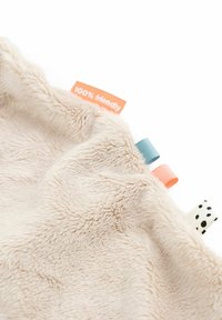 Tejido suave de felpa color beige con lazos de cinta coloridos y estampados, y una etiqueta naranja parcialmente visible que dice "100% amigable".