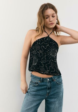 Mladá žena s vlnitými vlasmi má na sebe čierny halter top so striebornými vzormi a modré džínsy, pózuje s jednou rukou na krku.