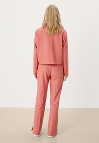 Roze cropped blazer en bijpassende broek, gladde stof, getailleerde pasvorm, rugsplitdetails, gedragen met witte sneakers op een beige vloer.