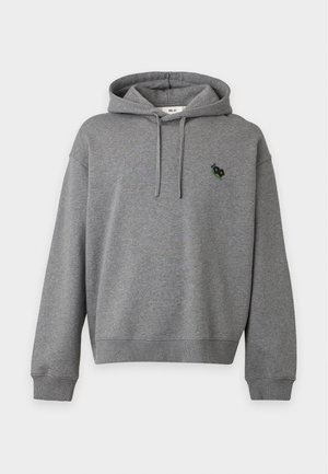 Jeune homme portant un sweat à capuche gris, un jean noir, des baskets grises et une casquette gris foncé, debout avec une main dans la poche devant un fond uni.