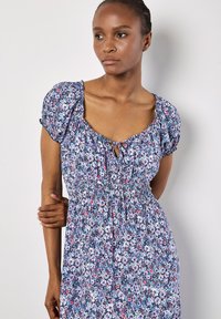 Blumenkleid in Blautönen mit Puffärmeln, einem quadratischen Ausschnitt und einer gerafften Taille. Schlüsselaugenakzent an der Vorderseite, leichtes Material.