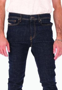 Homme portant un jean en denim bleu foncé et une chemise blanche, montrant la taille frontale, les poches et les mains reposant sur les côtés.