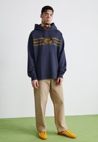 Tengerészeti kapucnis pulóver sárga csíkos grafikai designnal és logóval, bézs nadrággal és sárga tornacipővel párosítva. Az anyag pamut keveréknek tűnik.